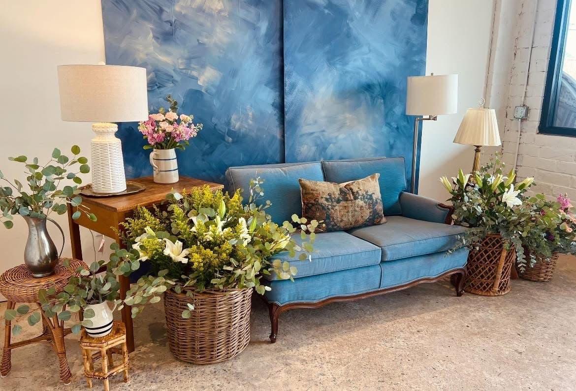 A styled corner of Tanglewood Arts Center with a blue sofa, artwork and flowers